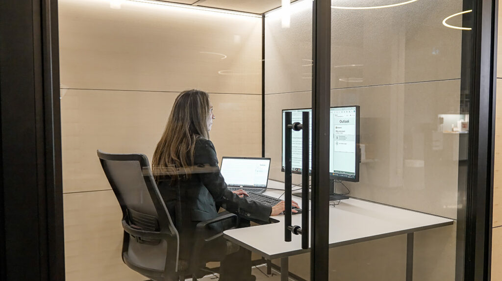Woman in a glass-walled office cubicle working at a desk with two monitors and a laptop.