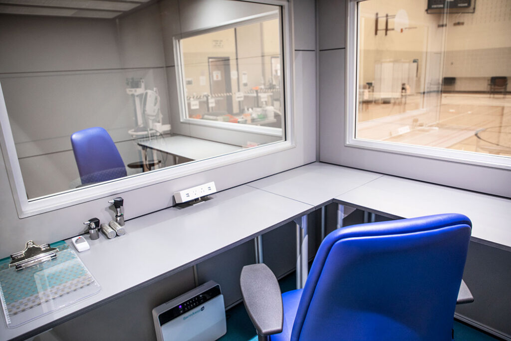 Observation booth with a blue chair and desk, looking through glass walls into a gym area