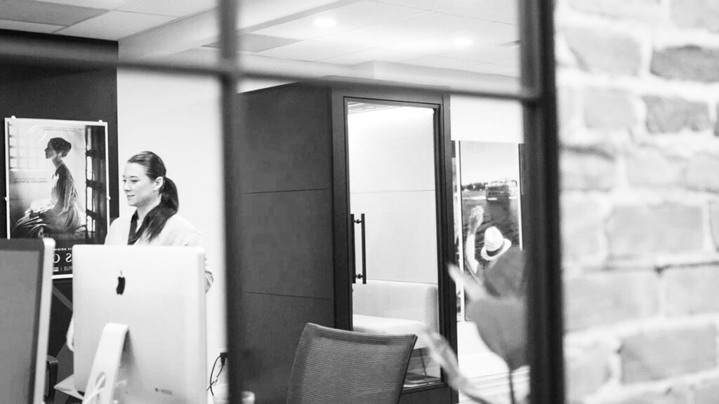 Office reception: woman in white blouse standing by a desk with an iMac, poster on the wall, glass partition and brick wall visible to the right.