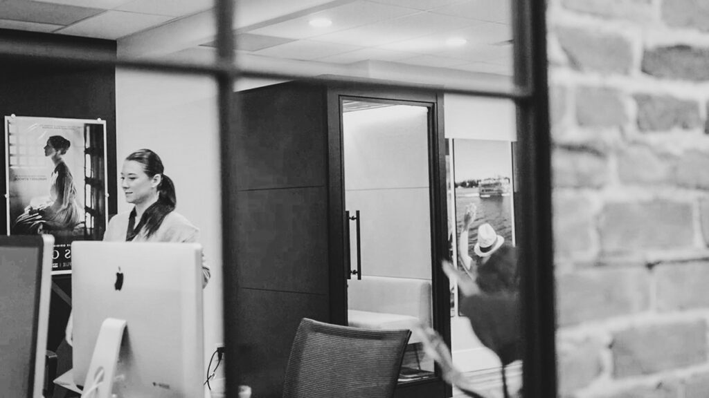Receptionist standing by an iMac at a modern office reception with a doorway and glass partition in the background.