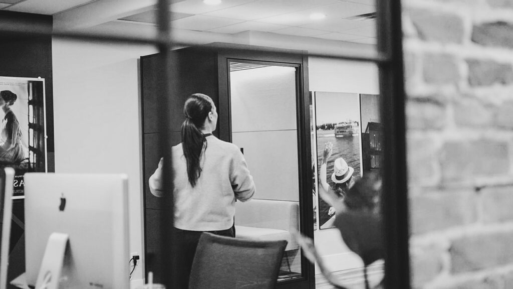 Black-and-white office scene: a woman with a ponytail enters a glass-walled meeting room.
