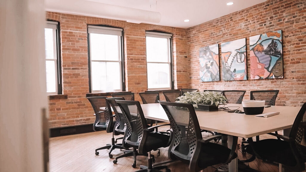 Bright brick-walled conference room with a long table, wheeled chairs, and three-panel mural on the wall.