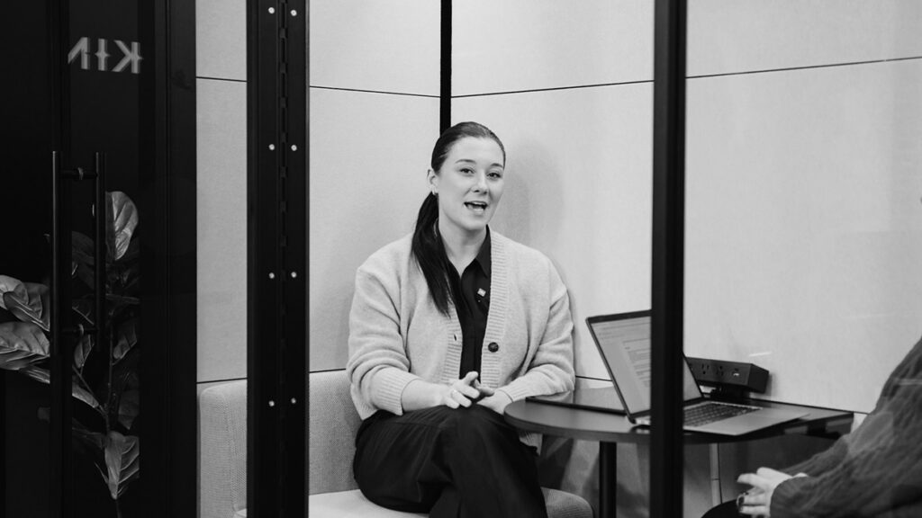 Woman in a light cardigan seated on a sofa inside a glass-walled meeting room, speaking with hands clasped, laptop on the round table beside her.