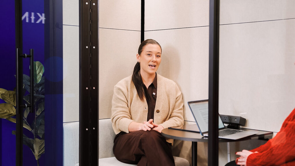 Woman seated in a glass-walled meeting pod, speaking with hands clasped, laptop on the round table beside her.