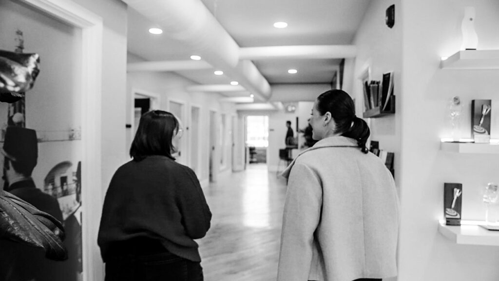 Two people walk away from the camera down a bright, modern hallway with items on shelves to the right.