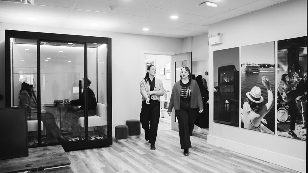 Two women walk through a modern office corridor; glass meeting room on the left and large wall photos on the right.