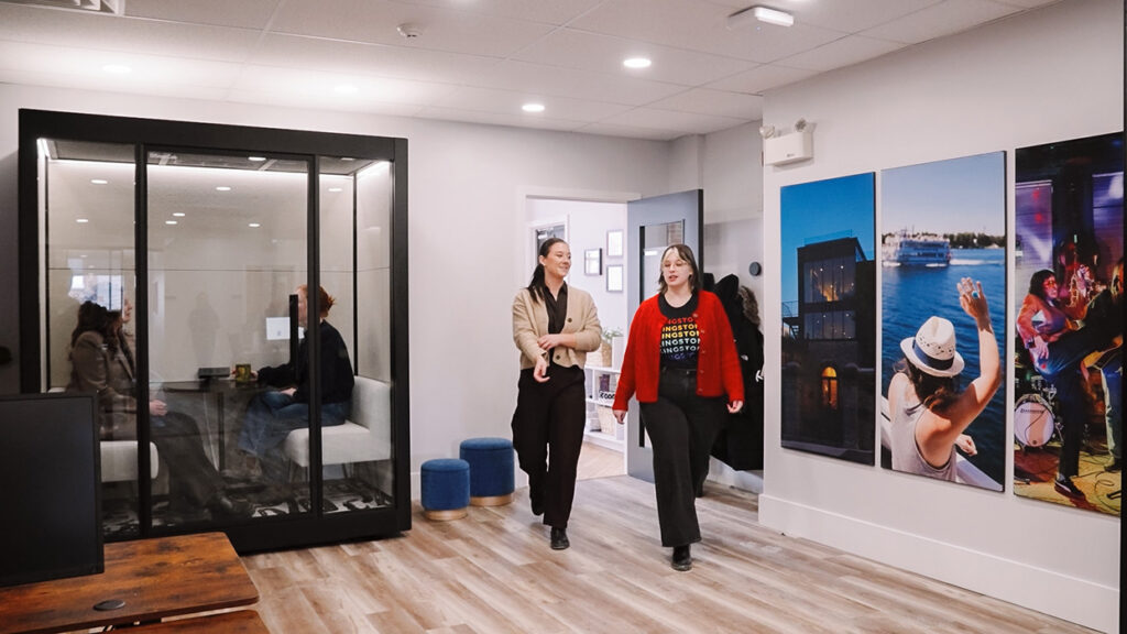 Two women walk down a bright office corridor with a glass‑enclosed meeting room on the left and large wall photos to the right.
