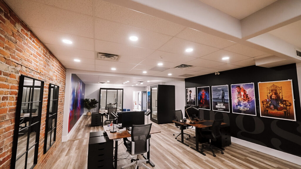 Open-plan office with an exposed brick wall on the left, workstations in the center, and movie posters on a black wall to the right.