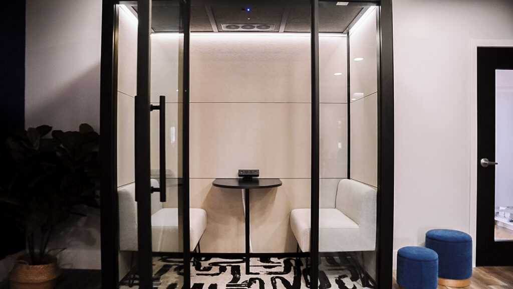 Glass-walled office pod with two white bench seats and a small round table, in a neutral lobby area.