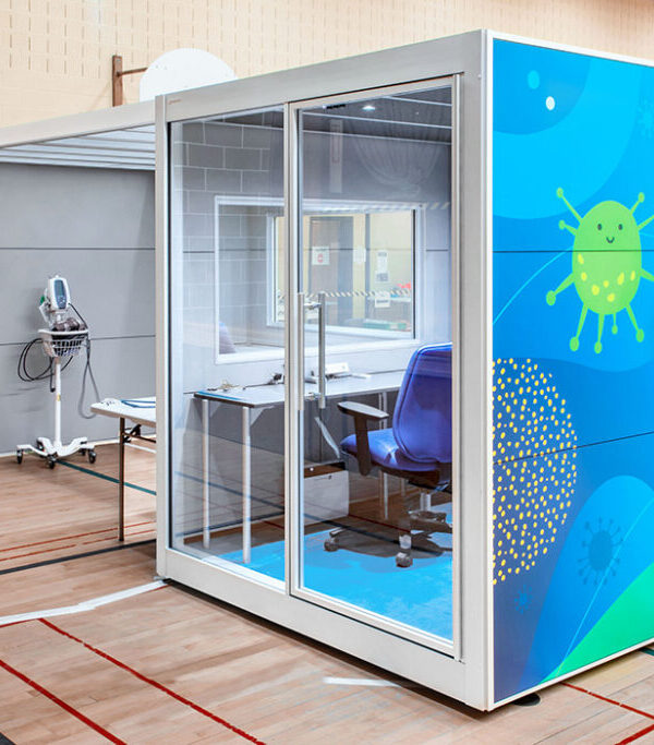 Medical testing booth in a gym, with a glass door and blue virus-themed exterior panel nearby surface medical equipment.