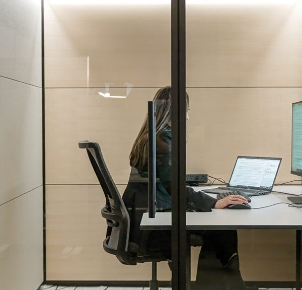 Person seated at a white desk inside a glass-walled private office, using a laptop and an external monitor.